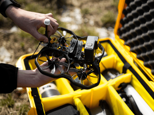Pilot holding a small FPV drone with mounted action camera above a yellow padded hard case containing aerial photography equipment.